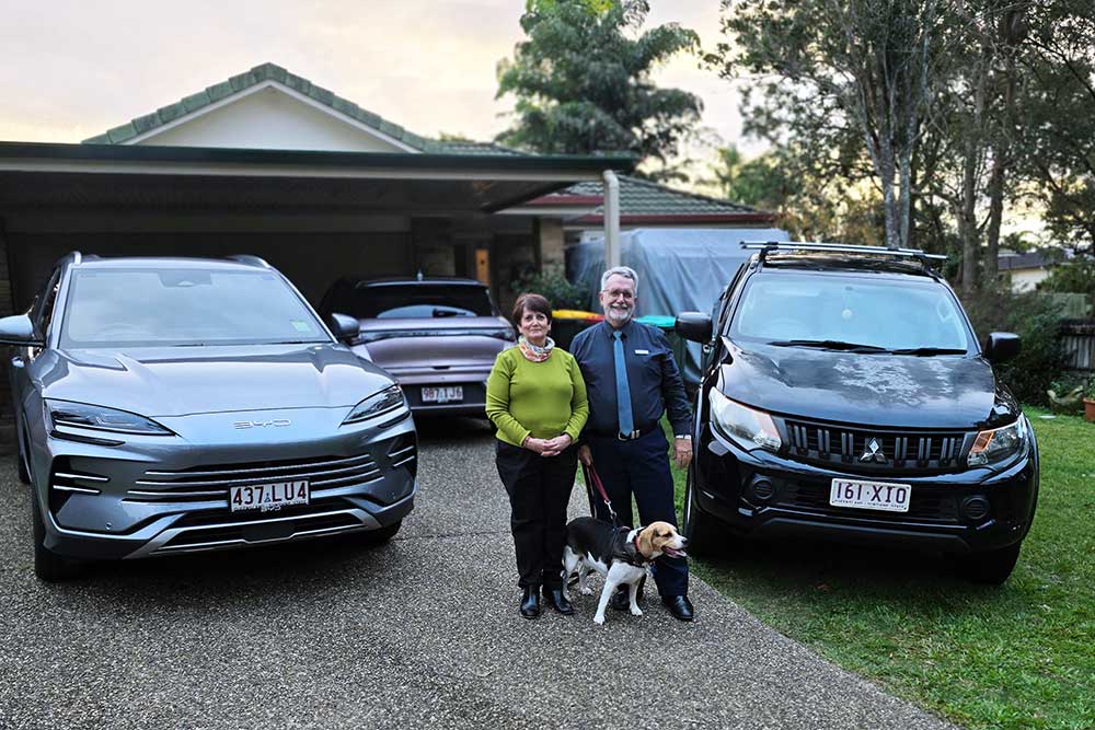 Husband and wife with their dog in front of their three cars.