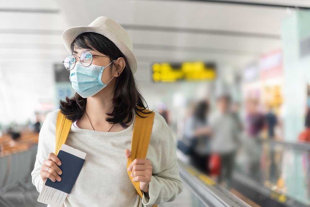 Woman walks through international airport holding passport and wearing a mask.