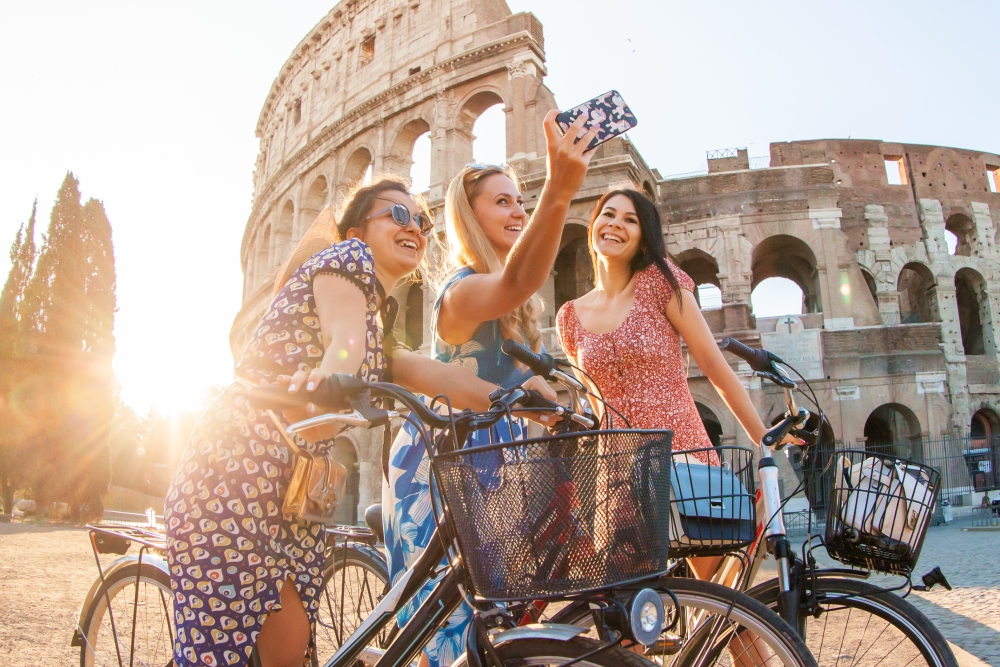 Three happy young women with bikes taking selfies at Colosseum in Rome, Italy