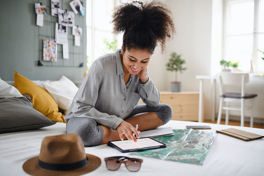 Woman sitting on a bed with her tablet and travel map smiling while planning