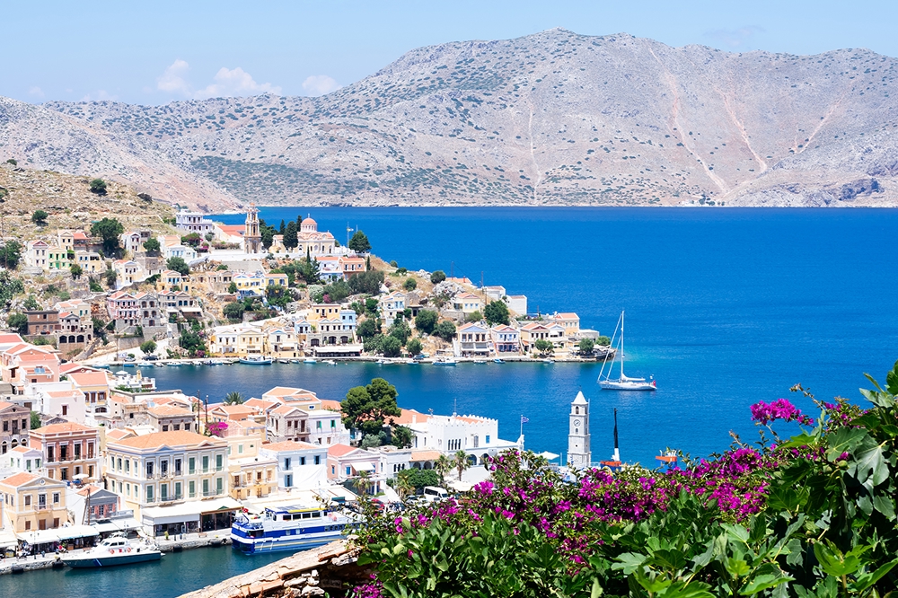 Picturesque view on tiny colourful houses on rocks and boats through blooming plants on balcony near the Mediterranian sea on Greek island