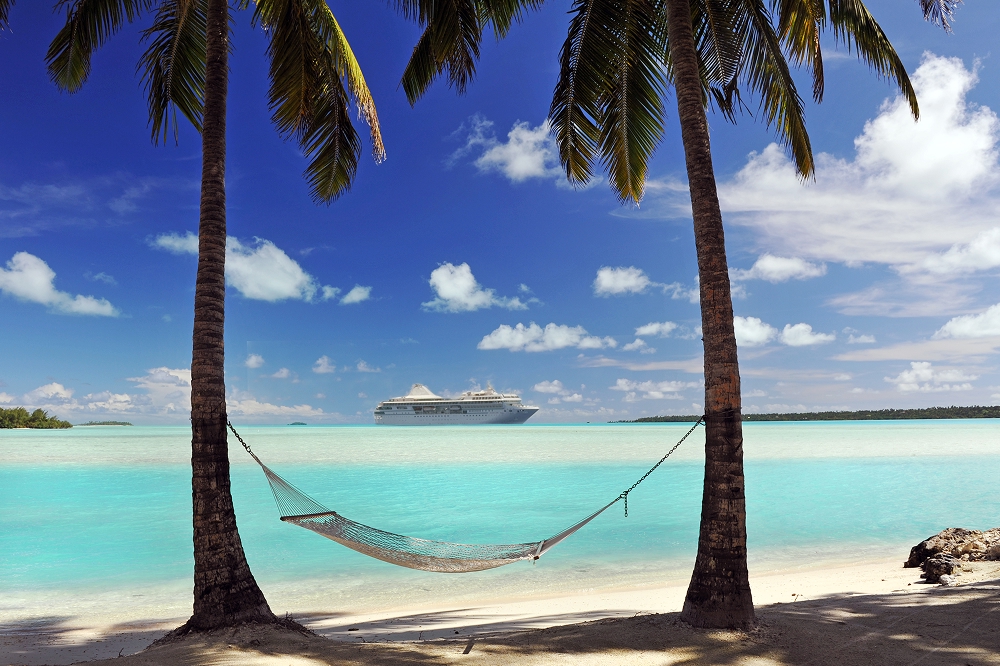 Closeup of hammock hung up between 2 palm trees on a tropical island, with a cruise ship sailing in the distance