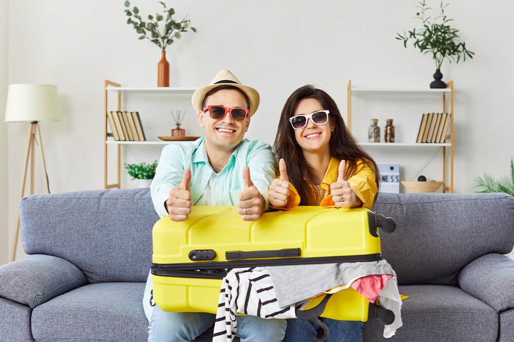 A young couple is preparing for their summer vacation sitting on the couch and showing thumbs up