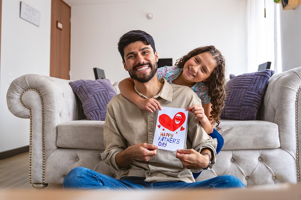 Child presenting a handmade Father’s Day card while sitting with their dad in a living room at home.