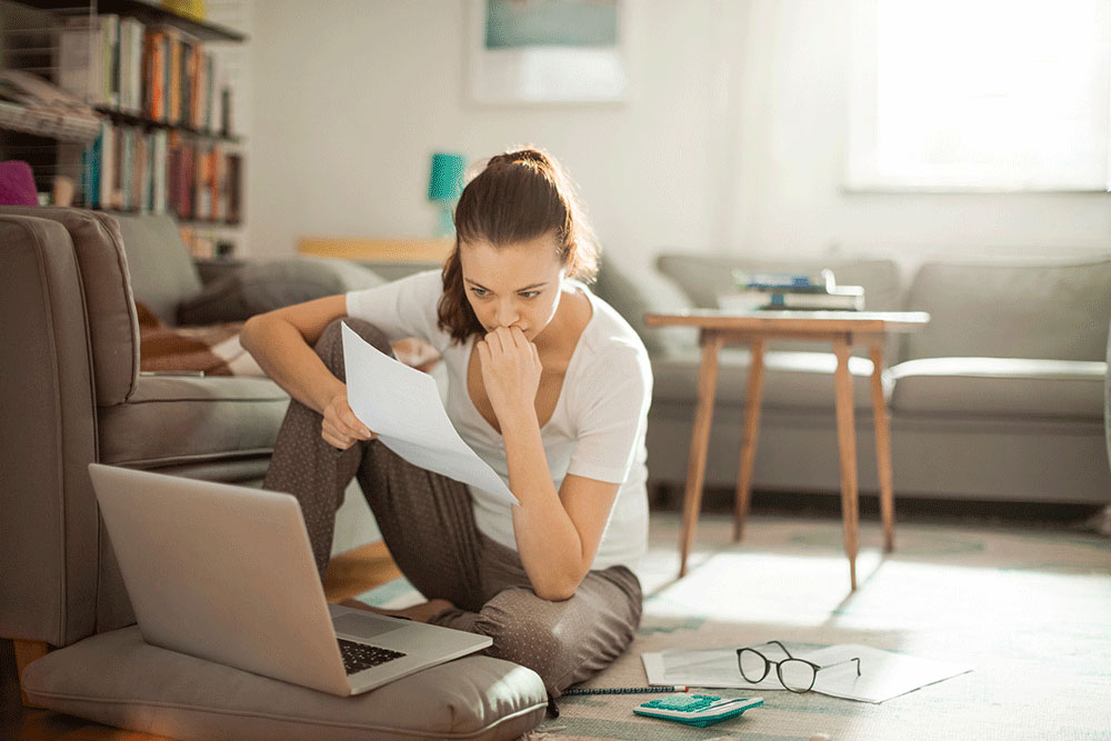 Woman sitting on the floor looking at a paper bill and a computer.