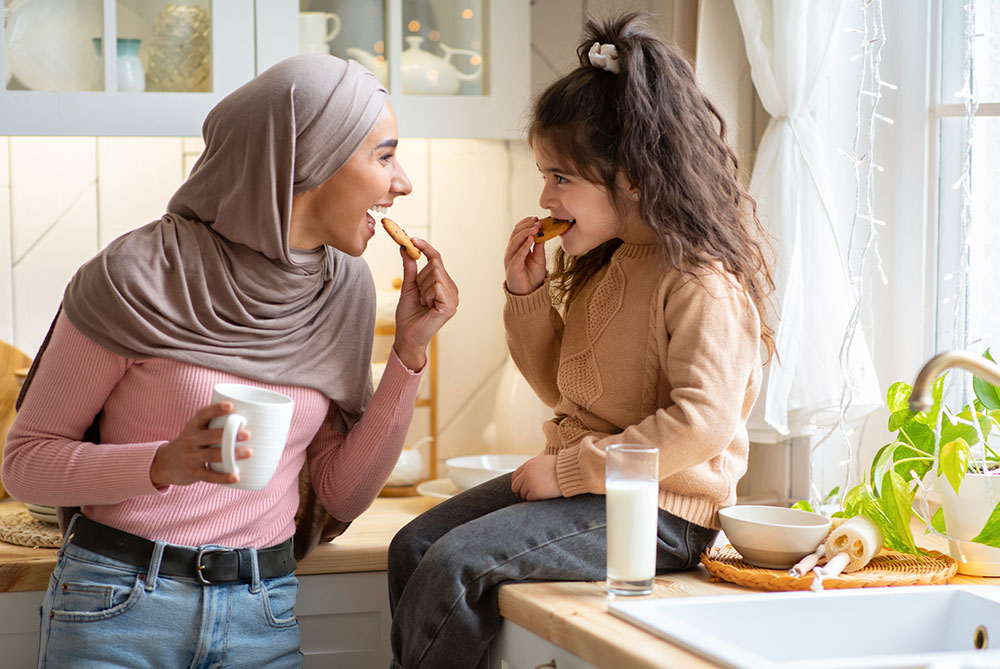 Muslim mum in hijab with coffee eating a cookie with daughter in the kitchen