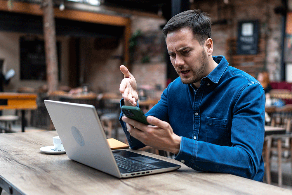 Man seems angry while looking at his phone and laptop.