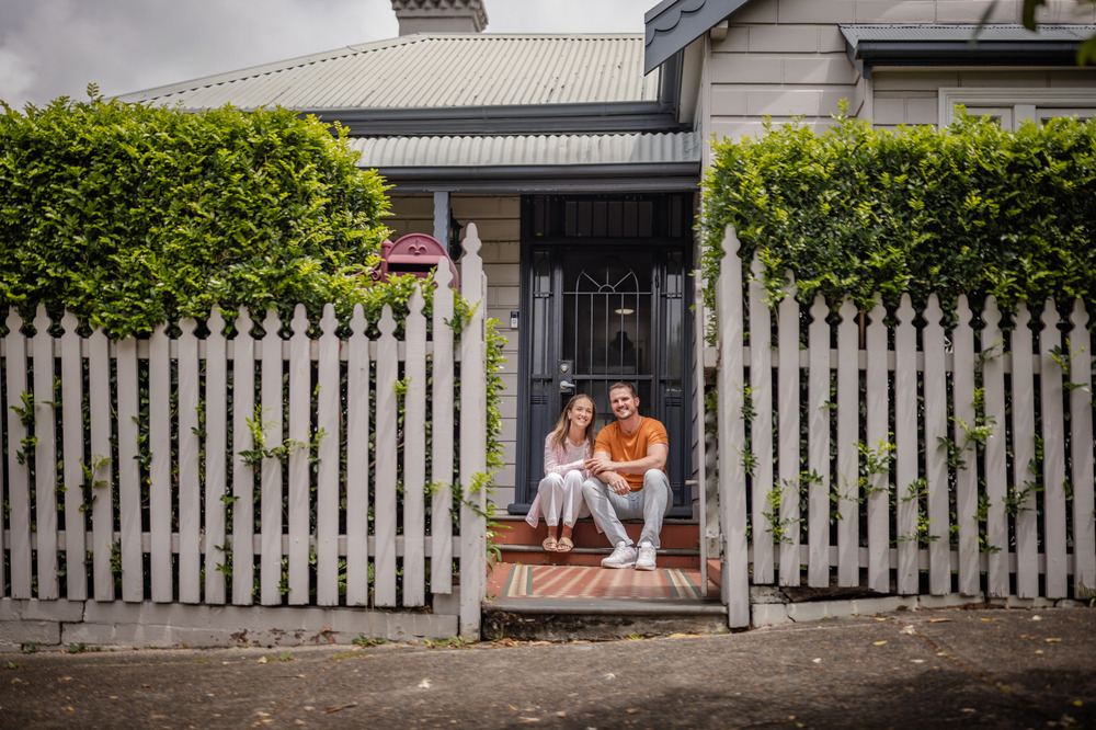 Young couple sitting infront of suburban home and white fence