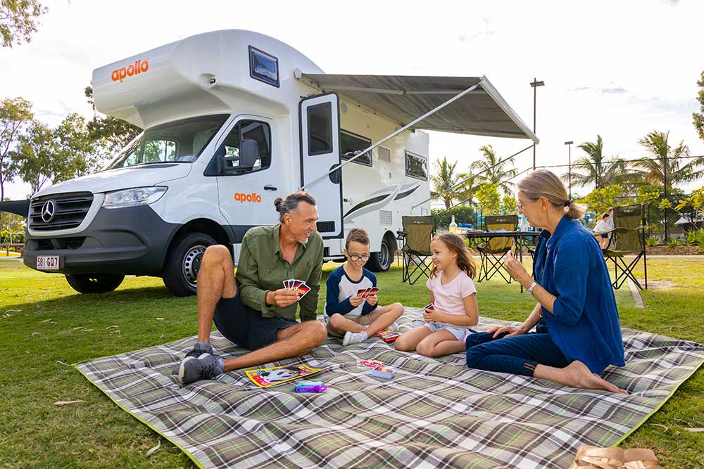 Family playing cards on picnic blanket beside campervan.