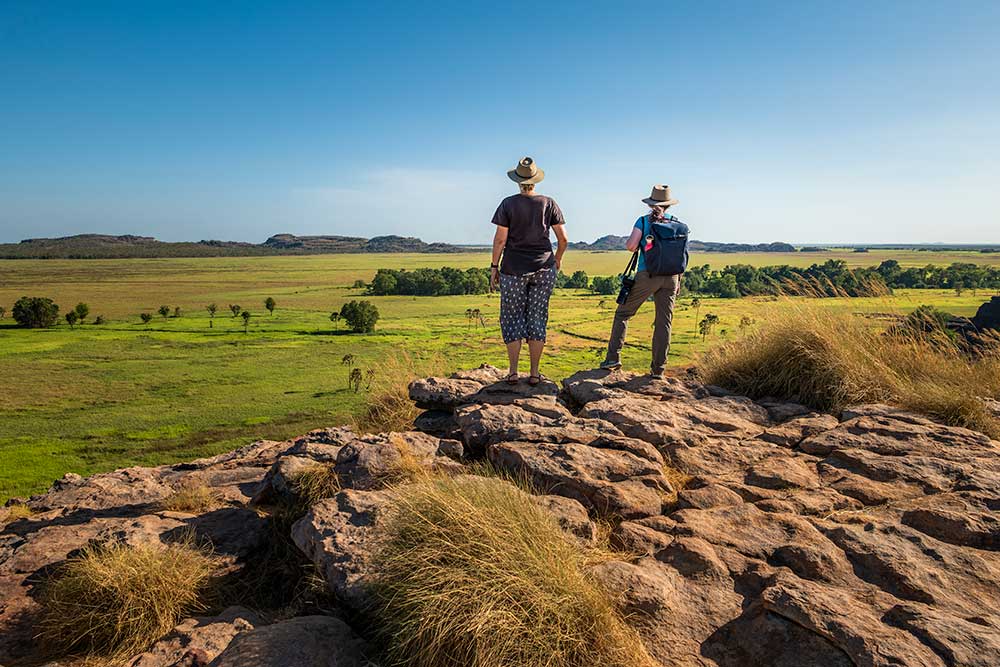 Two people looking at view across green plain from a rocky outcrop.