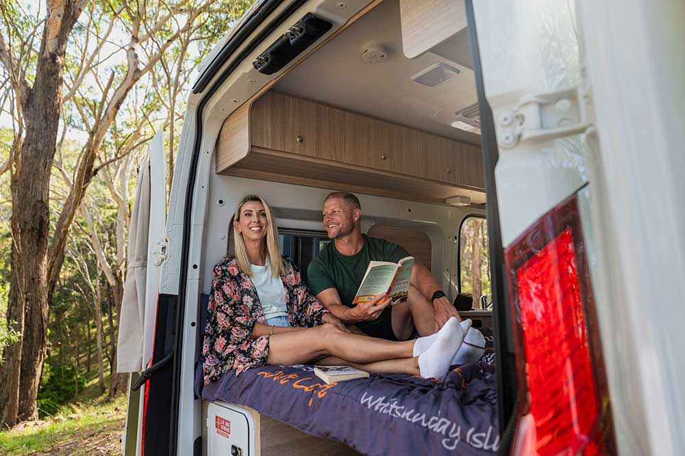 A woman and a man reading a book  in the back of a campervan.