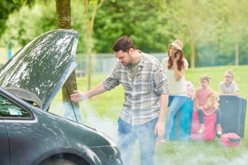 man assessing his broken-down car with front bonnet open and smoking while family behind him call roadside assistance