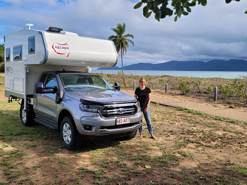 Woman standing beside a slide-on camper parked near a beach.