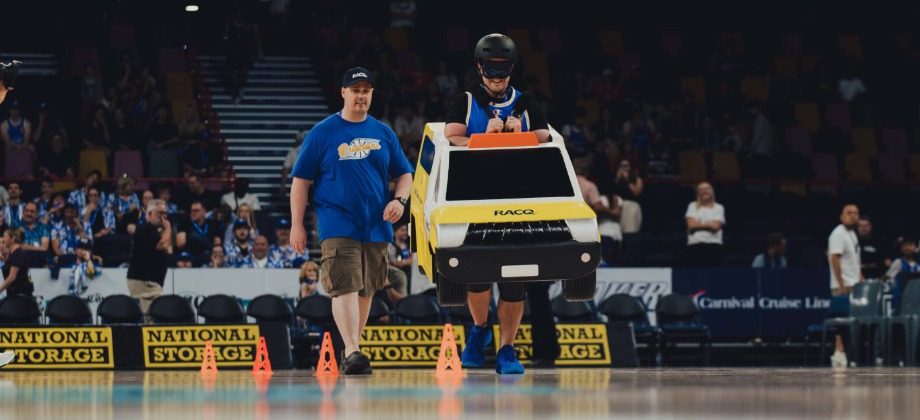 Person guiding a blindfolded person wearing an RACQ patrol car mascot costume on a basketball court