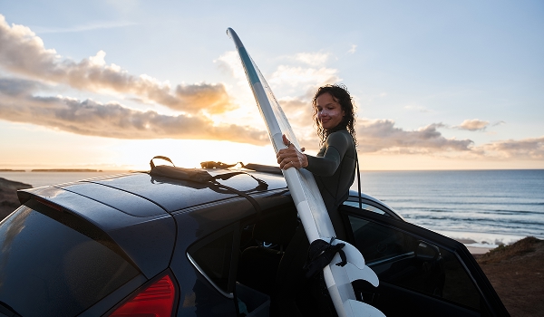 Person loading a surfboard onto a car beside the beach at sunset