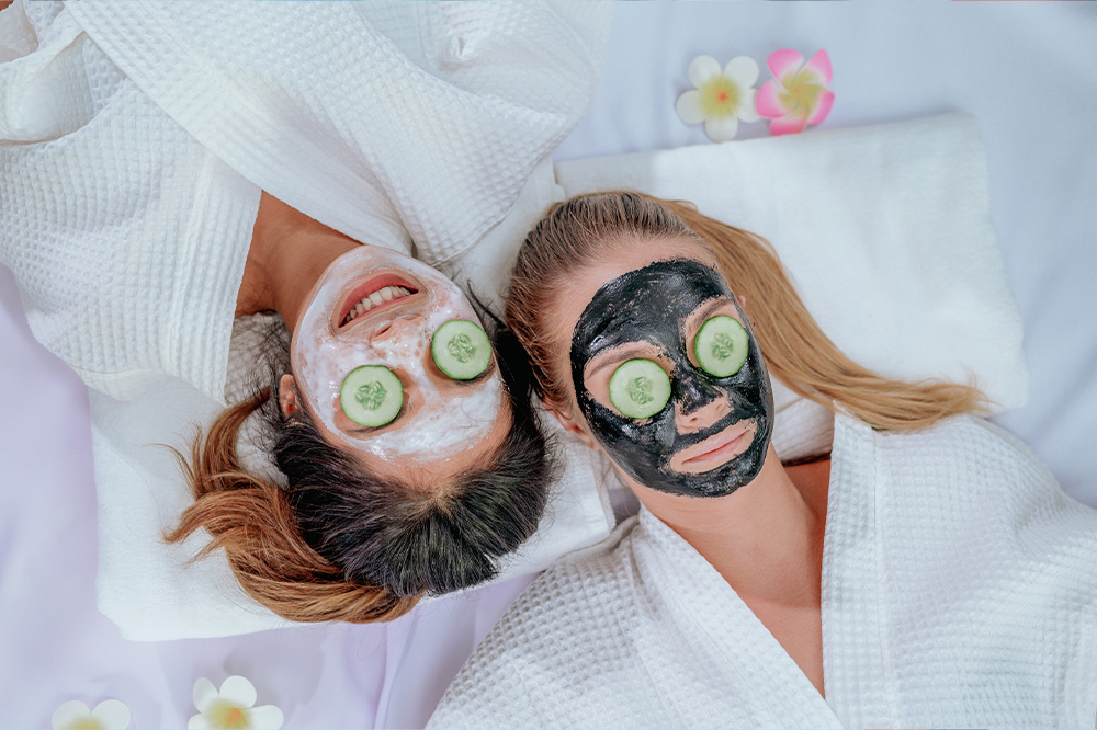 Two happy young women make cucumber or tomato facial skin care mask on eyes laughing relaxing together